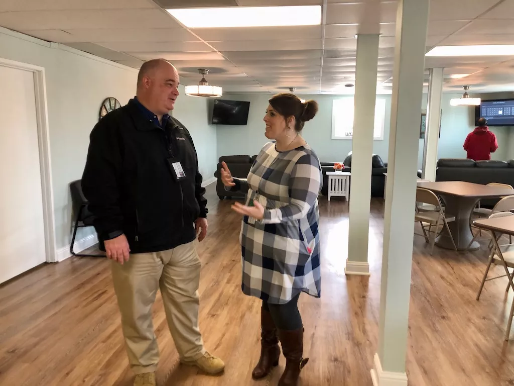 Two people standing and conversing inside an indoor common area with tables and seating in the background.